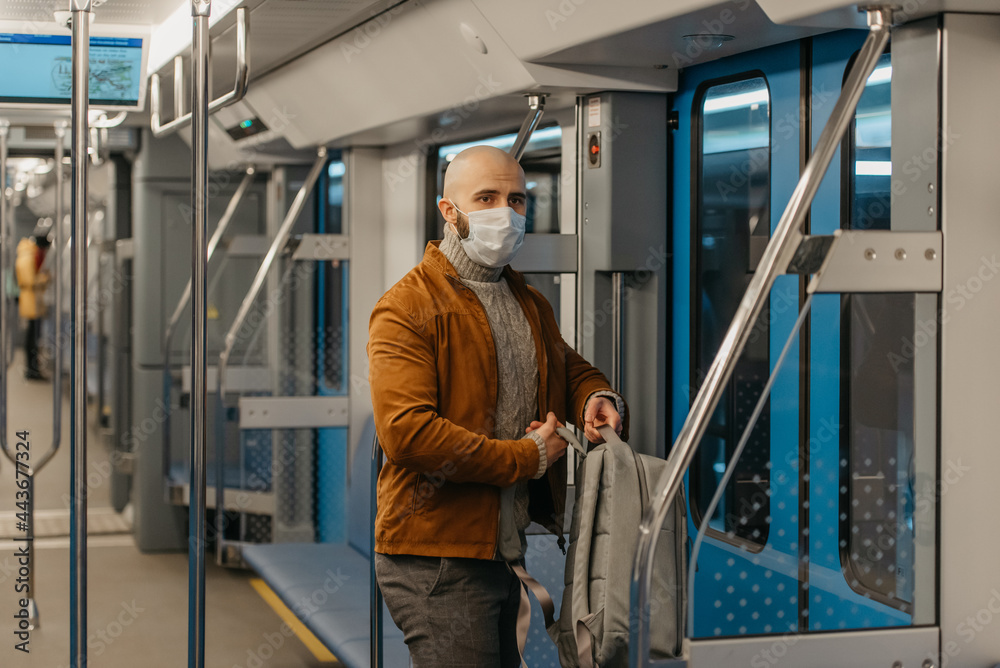 A man with a beard in a medical face mask to avoid the spread of coronavirus is putting on a backpack while riding a subway car. A bald guy in a surgical mask is keeping social distance on a train.