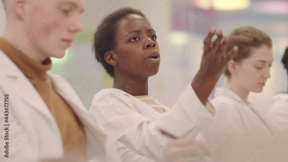 Medium side-view slowmo of young Afro American female medial student chatting with classmates during lecture asking them questions about new topic Medium side-view slowmo of young Afro American female medial student chatting with classmates during lecture asking them questions about new topic