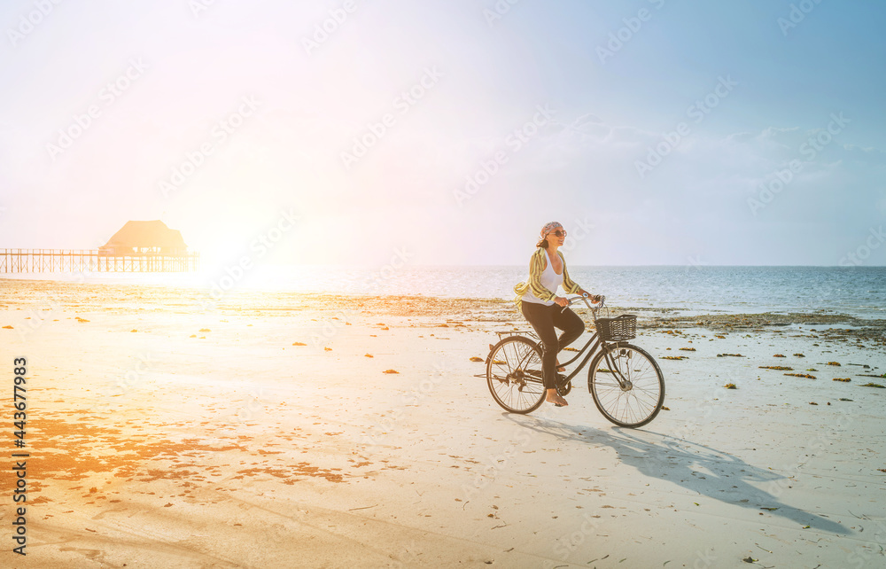 Obraz premium Young woman dressed light summer clothes riding old vintage bicycle with front basket on the lonely low tide ocean white sand coast on Kiwengwa beach on Zanzibar island, Tanzania.