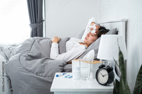 Asian woman feeling sick and sleeping in blanket on the bed while holding tissue in hand and touching forehead