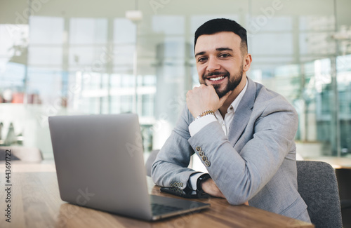 Cheerful businessman working on laptop and touching chin in office