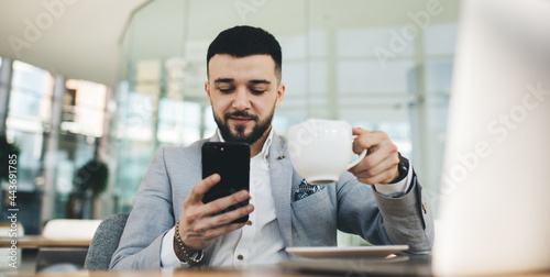 Positive businessman with smartphone and cup of coffee in office