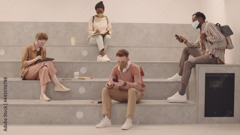 Wide shot with slowmo of group of young multiethnic students in face masks and casualwear sitting on stairs one by one scrolling on smartphones or taking notes in copybooks waiting for class