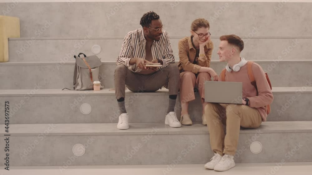 Wide shot of group of young multiethnic university students in casual clothes studying together on laptop preparing for project and chatting while sitting on stairs indoors