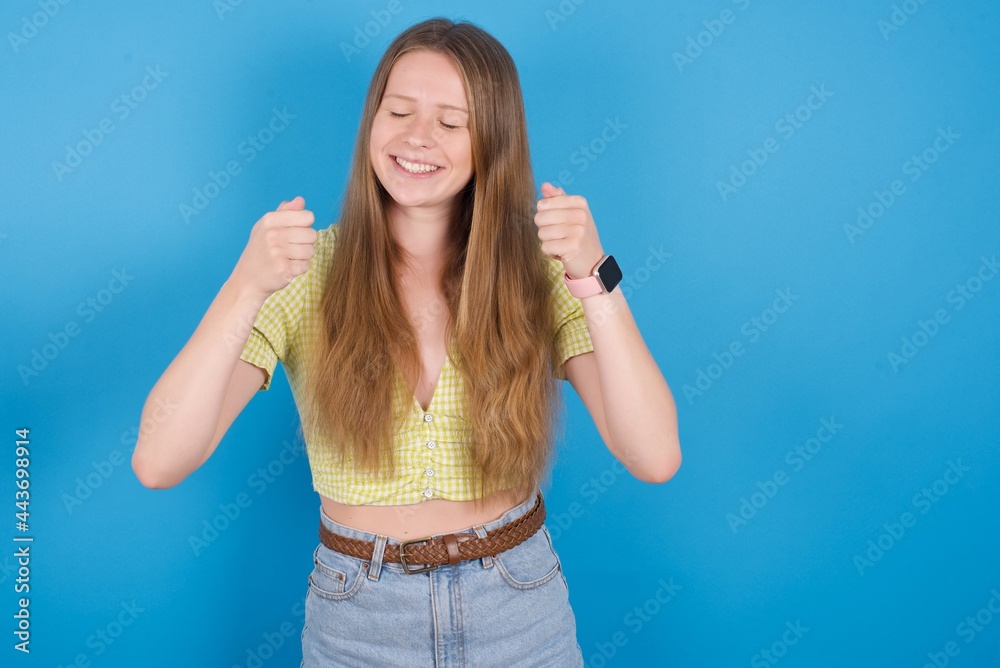 young beautiful blonde woman standing against blue background rejoicing his success and victory clenching fists with joy being happy to achieve aim and goals. Positive emotions, feelings. young beautiful blonde woman standing against blue background rejoicing his success and victory clenching fists with joy being happy to achieve aim and goals. Positive emotions, feelings.