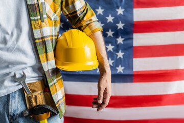 cropped view of workman in plaid shirt and tool belt holding hardhat near blurred usa flag, labor day concept