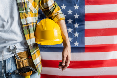 Bild auf Leinwand cropped view of workman in plaid shirt and tool belt holding hardhat near blurre
