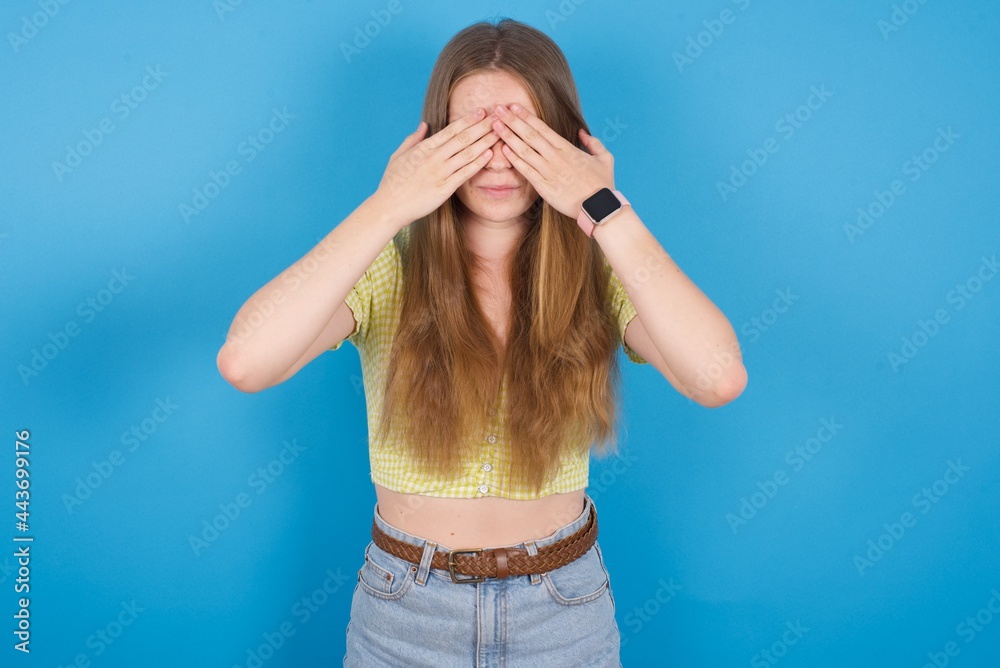 young beautiful blonde woman standing against blue background covering eyes with both hands, doesn't want to see anything or feeling ashamed. Human feelings reactions.