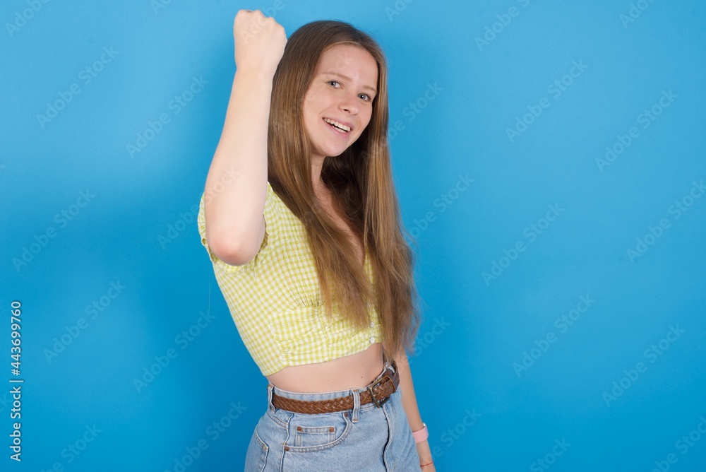 Overjoyed young beautiful blonde woman standing against blue background glad to receive good news, clenching fist and making winning gesture. Overjoyed young beautiful blonde woman standing against blue background glad to receive good news, clenching fist and making winning gesture.