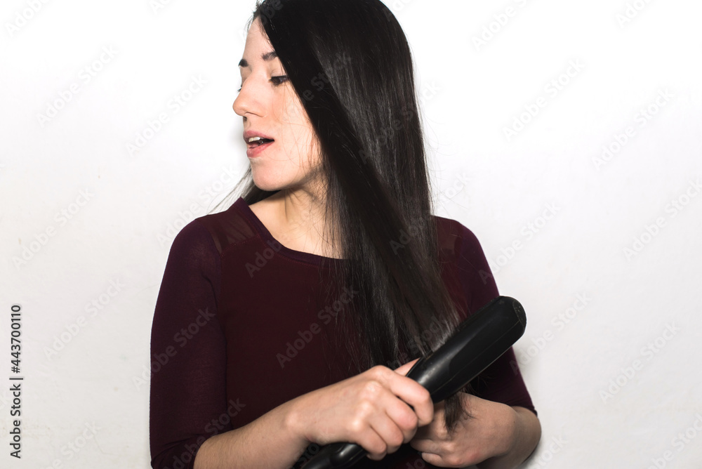 Smiling young woman straightening her long black shiny hair using a flat iron isolated on a white background. Hair straightener Smiling young woman straightening her long black shiny hair using a flat iron isolated on a white background. Hair straightener