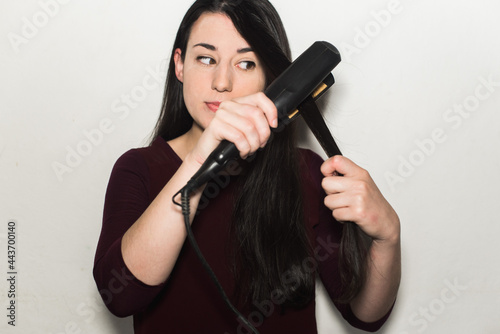 Pretty european young woman straightening her long black shiny hair using a flat iron isolated on a white background photo