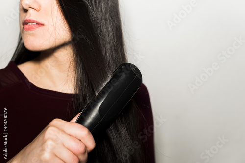 Close-up of a young woman straightening her long dark brown shiny hair with a flat iron isolated in a white background