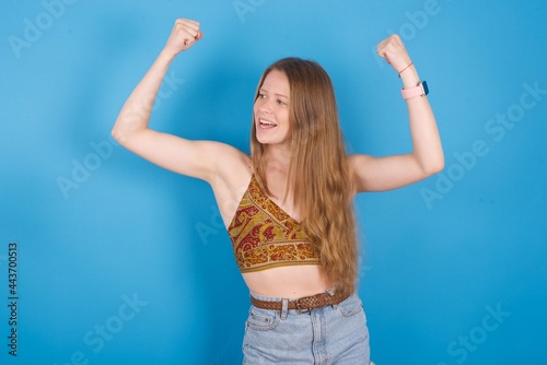 Yes I am winner. Portrait of charming delighted and excited young beautiful blonde woman standing against blue b raising up fist in triumph and victory smiling achieving success grinning from delight.