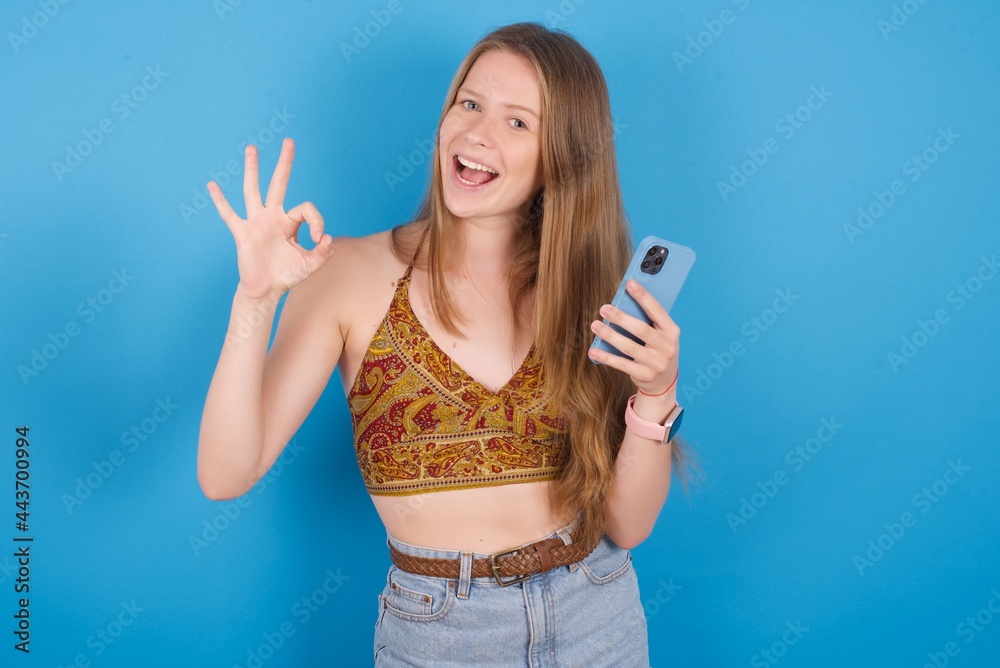 Happy young beautiful blonde woman standing against blue background sending a message on his smartphone or taking a selfie and making ok sign with his hand. Happy young beautiful blonde woman standing against blue background sending a message on his smartphone or taking a selfie and making ok sign with his hand.