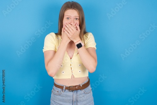 Stunned young beautiful blonde woman standing against blue background covers mouth with both hands being afraid from something or after hearing stunning gossips.