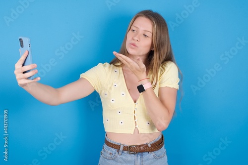 young beautiful blonde woman standing against blue background blows air kiss at camera of smartphone and takes selfie  sends mwah via online call.