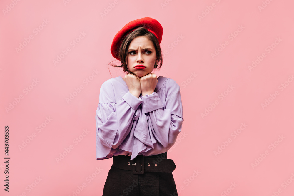Nice girl dressed in purple blouse, red beret and pants posing offendedly on pink background