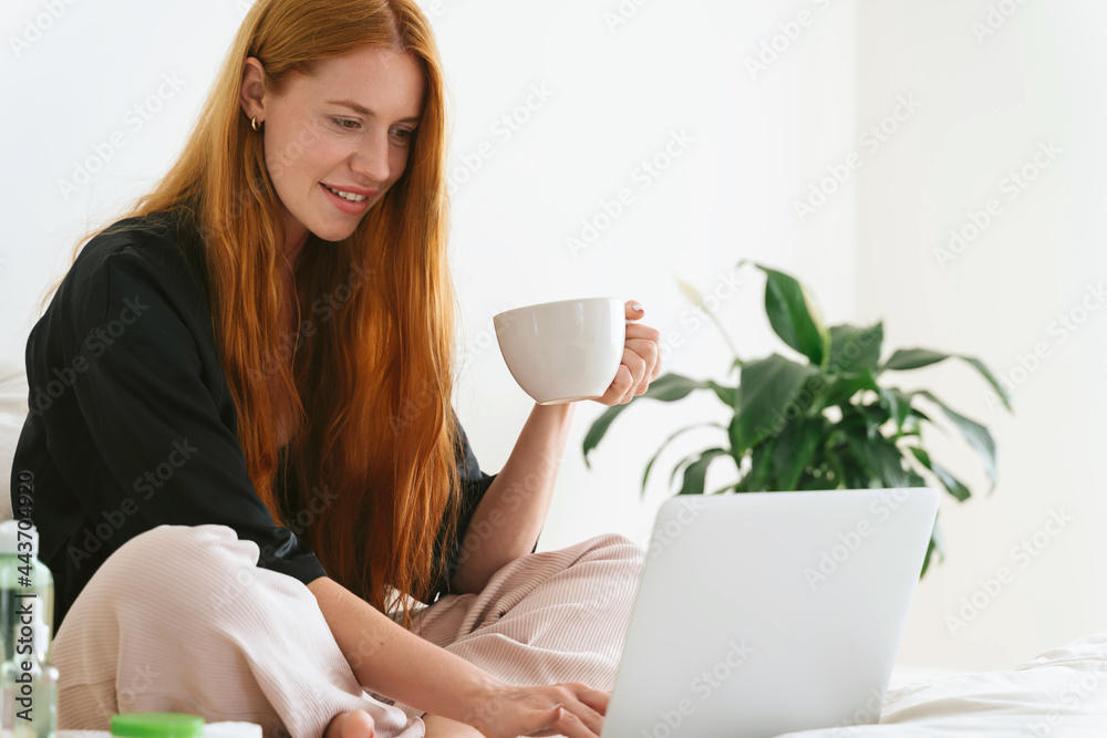 Red hair young girl working on laptop at home. Happy relaxed redhead beautiful woman using notebook in bedroom. Freelancer and remote worker doing his work concept. Red hair young girl working on laptop at home. Happy relaxed redhead beautiful woman using notebook in bedroom. Freelancer and remote worker doing his work concept.