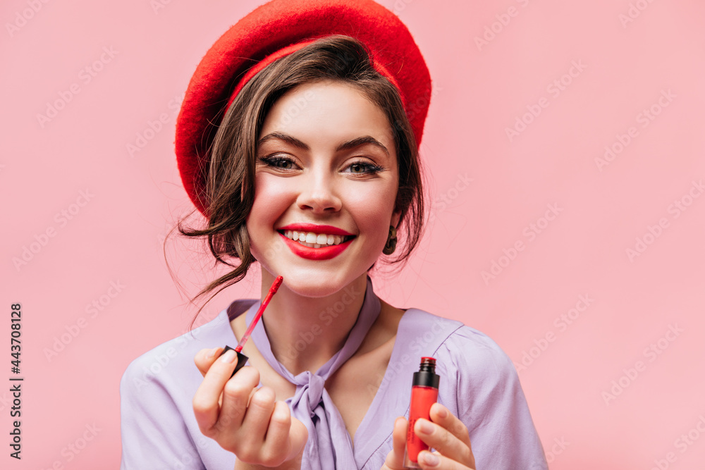 Green-eyed girl with snow-white smile paints her lips with red lipstick. Portrait of lady in stylish beret on pink background Green-eyed girl with snow-white smile paints her lips with red lipstick. Portrait of lady in stylish beret on pink background