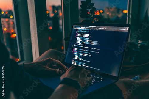 The code is on a laptop on a wooden table in front of the window  in the dark with a view of the lights of the night city, color lighting in the room, home decor	 photo