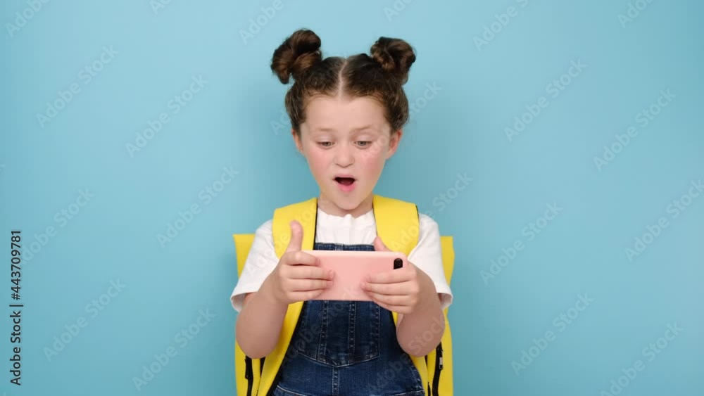 Smiling schoolgirl with backpack holding smartphone enjoying using mobile apps, playing video games, child learning in cellphone, having fun with mobile technology concept, posing on blue background Smiling schoolgirl with backpack holding smartphone enjoying using mobile apps, playing video games, child learning in cellphone, having fun with mobile technology concept, posing on blue background