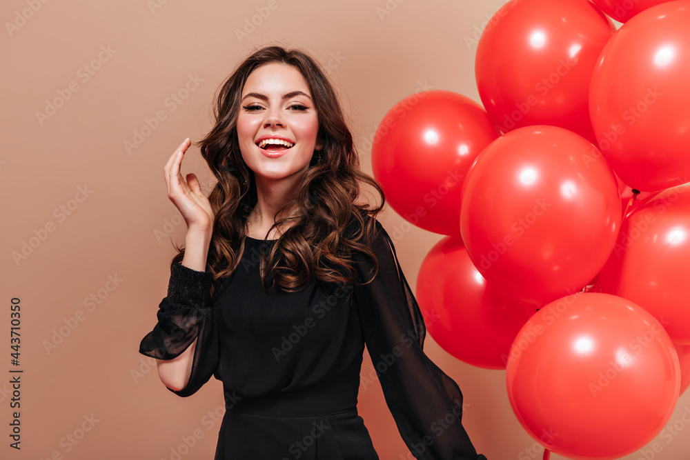 Nice girl in black light blouse with smile looks into camera and poses on beige background with balloons Nice girl in black light blouse with smile looks into camera and poses on beige background with balloons