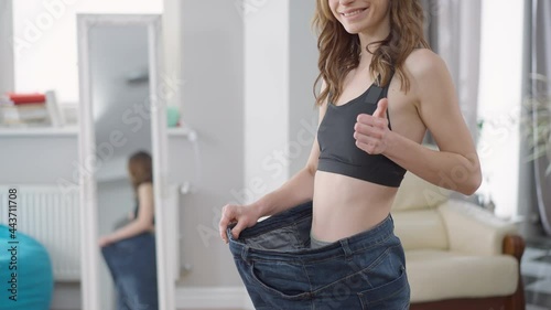 Unrecognizable slim young woman in oversize jeans showing thumb up smiling. Slender fit Caucasian millennial showing off training and dieting result standing at home indoors. Satisfaction and beauty photo