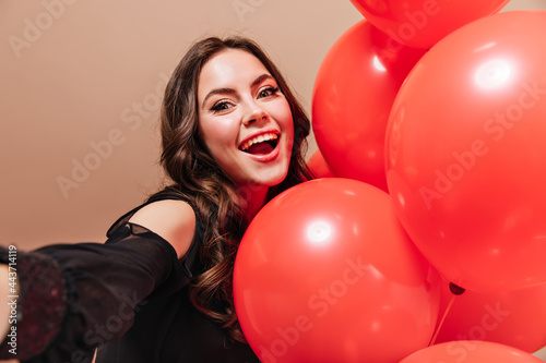 Cheerful curly lady smiles, looks into camera and takes selfie with balloons