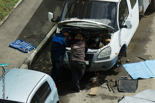 Two men are repairing a truck in the yard. The wheel has been removed from the car and the trunk is open. Tools are lying around. Auto mechanics stand at the open hood.