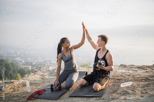 Happy multiracial couple in sportswear giving high five to each other while sitting on yoga mat outdoors. Black woman and caucasian man training with sport equipments.