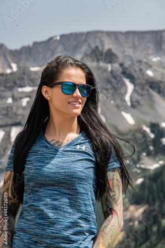 Portrait of a young brunette woman with tattoos  smiling with a mountain behind her. She is tan and has blue sunglasses on. 