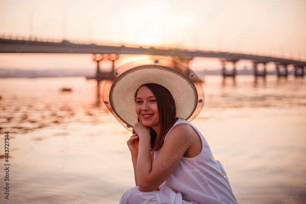 Fototapeta premium a laughing, charming lady in an elegant hat and a white summer dress is having a good summer evening on the beach under the bridge by the river. 
