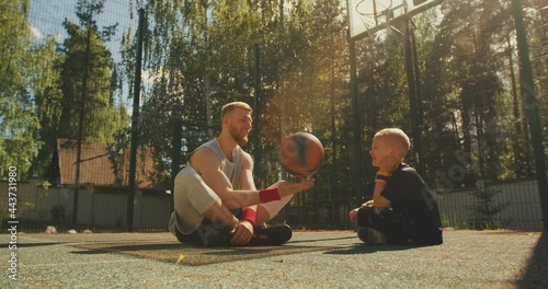 Sports happy family. Male basketball player spinning a ball on finger sitting with boy on basketball court