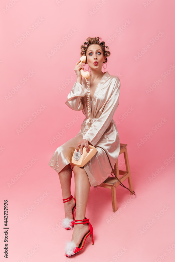 Girl with curlers dressed in robe sits on chair and speaks in surprise on phone. Girl with curlers dressed in robe sits on chair and speaks in surprise on phone.