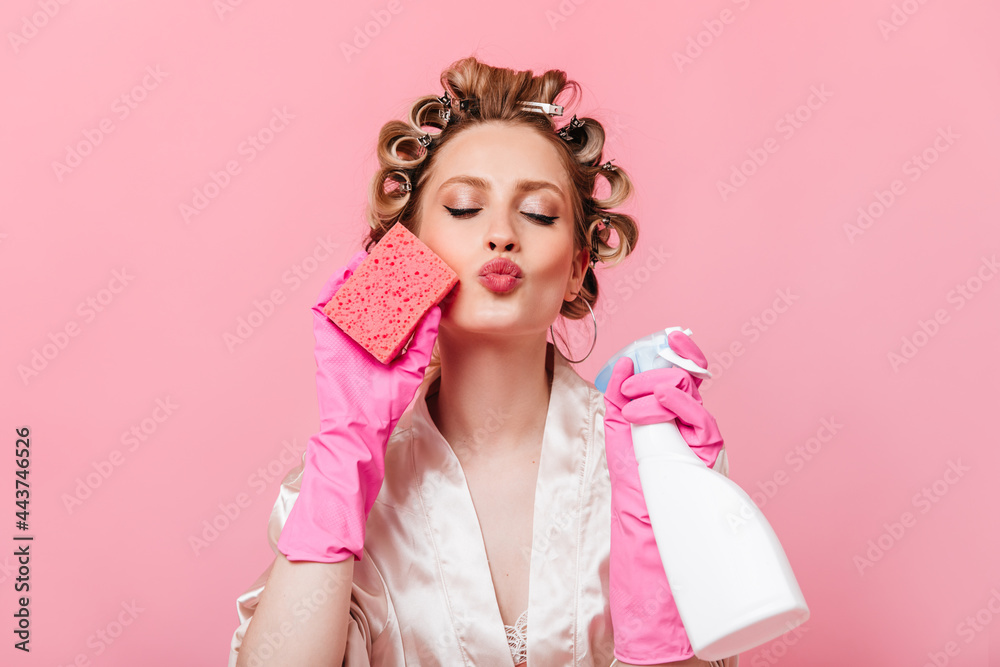 Housewife in pink robe and rubber gloves sends kiss. Girl holds instruments for cleaning Housewife in pink robe and rubber gloves sends kiss. Girl holds instruments for cleaning