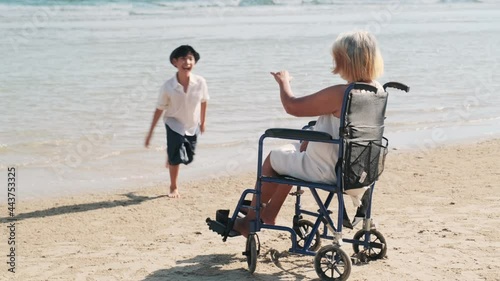 Elderly Asian woman sits in a wheelchair on the beach, talking and smiling with her grandson.