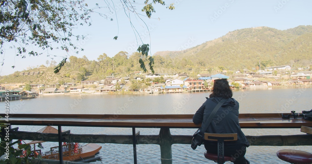 Obraz premium Rear view Young Asian tourist traveler woman sitting relaxing enjoy vacation looking at nature pond lake with village and hill on background at Ban Rak Thai village, Mae hong son, Northern Thailand 