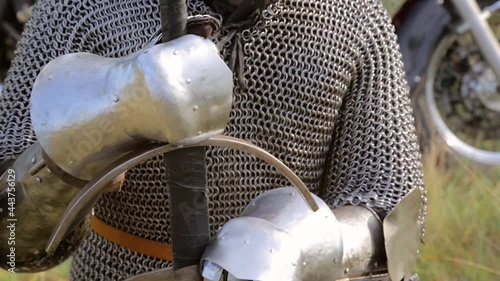 A knight in a full set of medieval armor and with a bandana on his head sits on one knee with a two-handed sword in his hands in a pose for prayer.