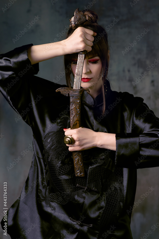 Beautiful woman with bright make up posing in Chinese costume in minimalistic interior with fan and dagger