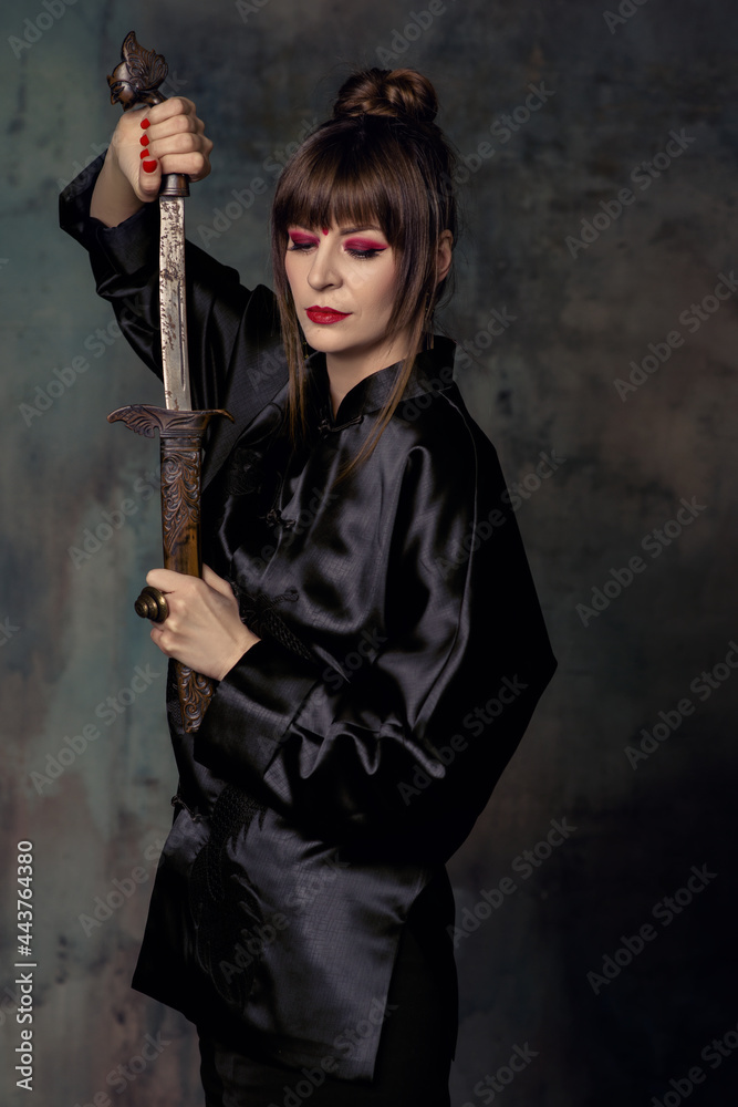 Beautiful woman with bright make up posing in Chinese costume in minimalistic interior with fan and dagger Beautiful woman with bright make up posing in Chinese costume in minimalistic interior with fan and dagger