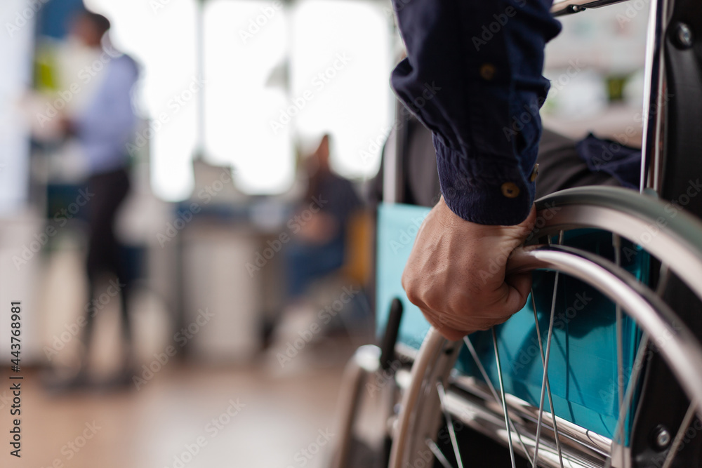 Fototapeta premium Close up independence handicapped man in wheelchair entering in company office, moving alone starting to work at financial project using professional computer after greeting colleagues.