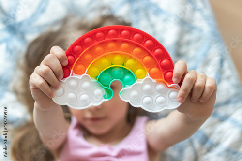 Little blonde girl holds out a toy pop it in the shape of a rainbow in her hands