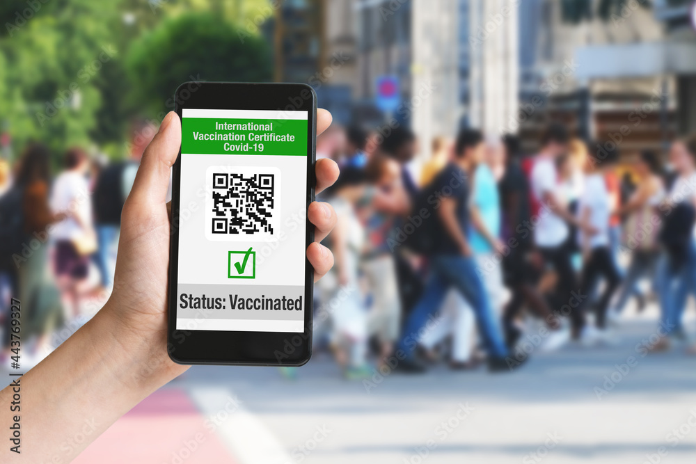 Hand holding mobile phone in city with international vaccination certificate on display as symbol for immunization Hand holding mobile phone in city with international vaccination certificate on display as symbol for immunization
