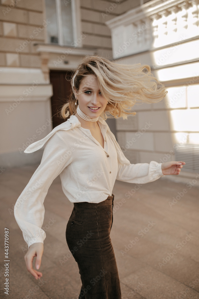 Grey-eyed curly blonde woman in brown pants and white blouse smiles, looks into camera, turns and walks outside. Grey-eyed curly blonde woman in brown pants and white blouse smiles, looks into camera, turns and walks outside.