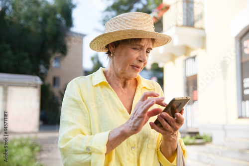 happy senior woman in sun hat  using smartphone, looking at screen on summer city