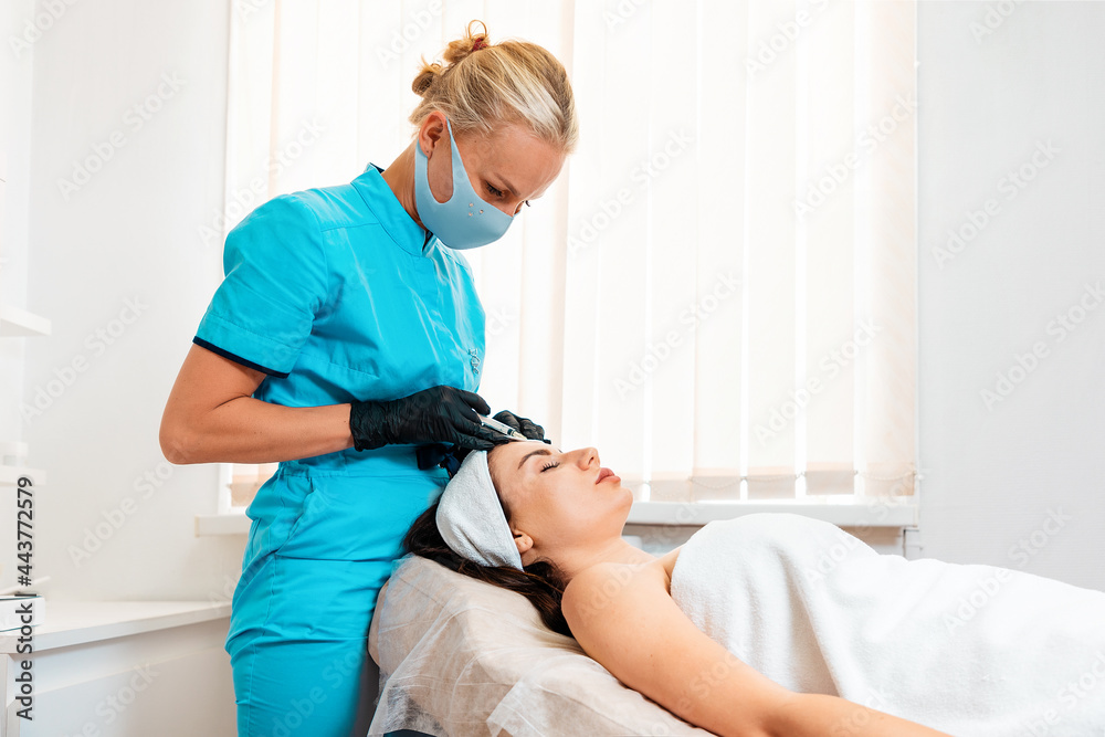 Woman getting injection at her forehead in the cosmetology salon. Cosmetologist in latex gloves and protective mask makes with syringe injects of a hyaluronic acid. Concept of plastic surgery Woman getting injection at her forehead in the cosmetology salon. Cosmetologist in latex gloves and protective mask makes with syringe injects of a hyaluronic acid. Concept of plastic surgery