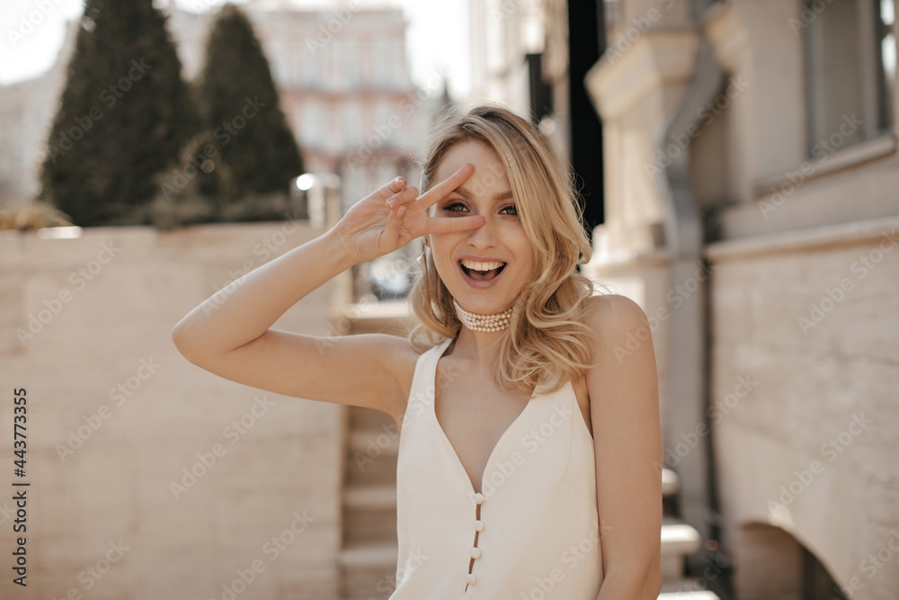 Fototapeta premium Pretty blonde attractive woman in pearl necklace and white silk dress shows peace sign, looks into camera and smiles outside.