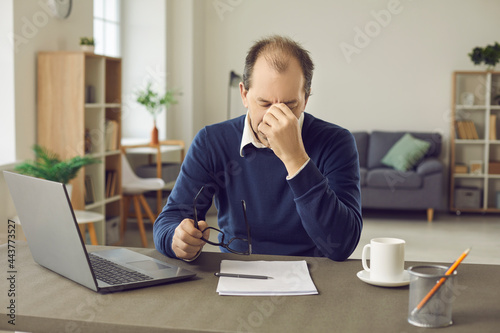 Senior business man who is tired from the workload suffers from headaches and lack of sleep. Exhausted male employee wipes his eyes suffering from unproductiveness and burnout. Concept of fatigue.