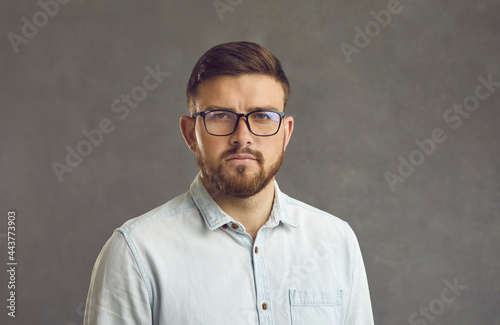 Close up studio portrait of millennial man with serious facial expression on gray background. Bearded man in glasses and a denim shirt looks straight into the camera with a simple and natural look.