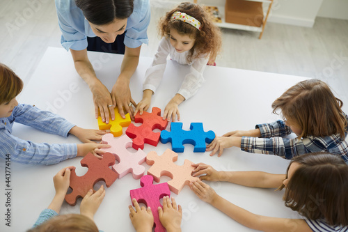Female teacher engaging kids in teamwork. Little children participate in fun group activity in class. Small team of active students together joining big colorful jigsaw puzzle parts on classroom table